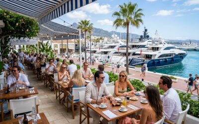 Outdoor café terrace in Puerto Banús with people socialising, yachts in the marina, palm trees, and shops along the promenade on a sunny day.